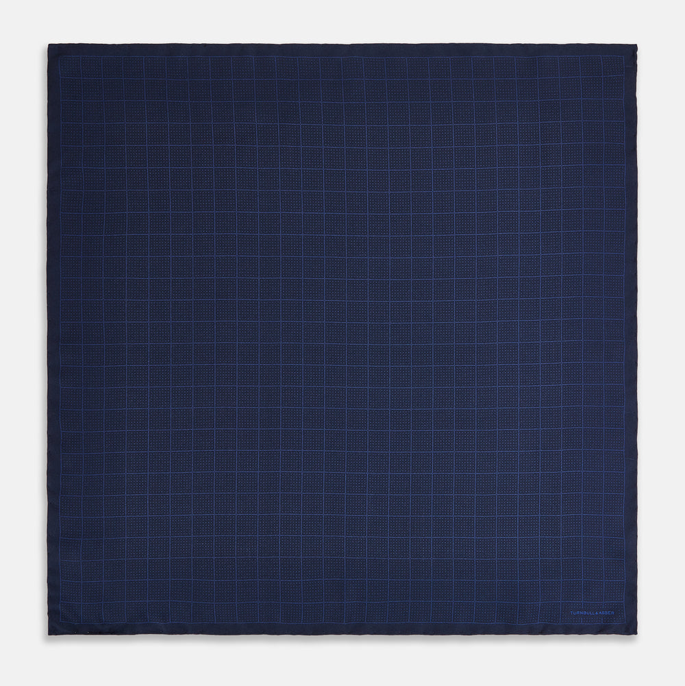 The Checked Blue On Navy Pocket Square in Silk by Turnbull & Asser UK is crafted from dark blue silk and features a subtle geometric design with a lighter blue grid pattern, finished with a discreet Turnbull & Asser logo in the corner.