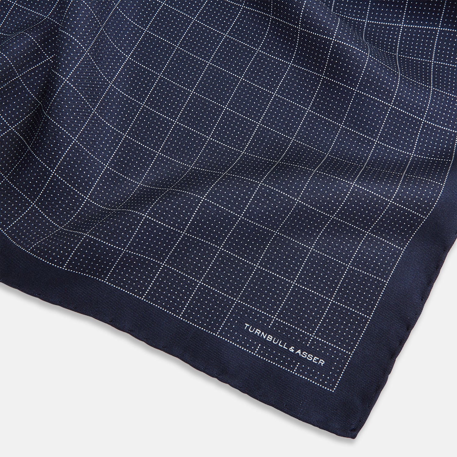 View 2 of product Checked Navy On White Pocket Square in Silk by Turnbull & Asser UK, featuring a navy blue geometric pattern with a white dotted grid and a solid border. The pocket square is shown with one corner folded over to display the “TURNBULL & ASSER” label in white text.