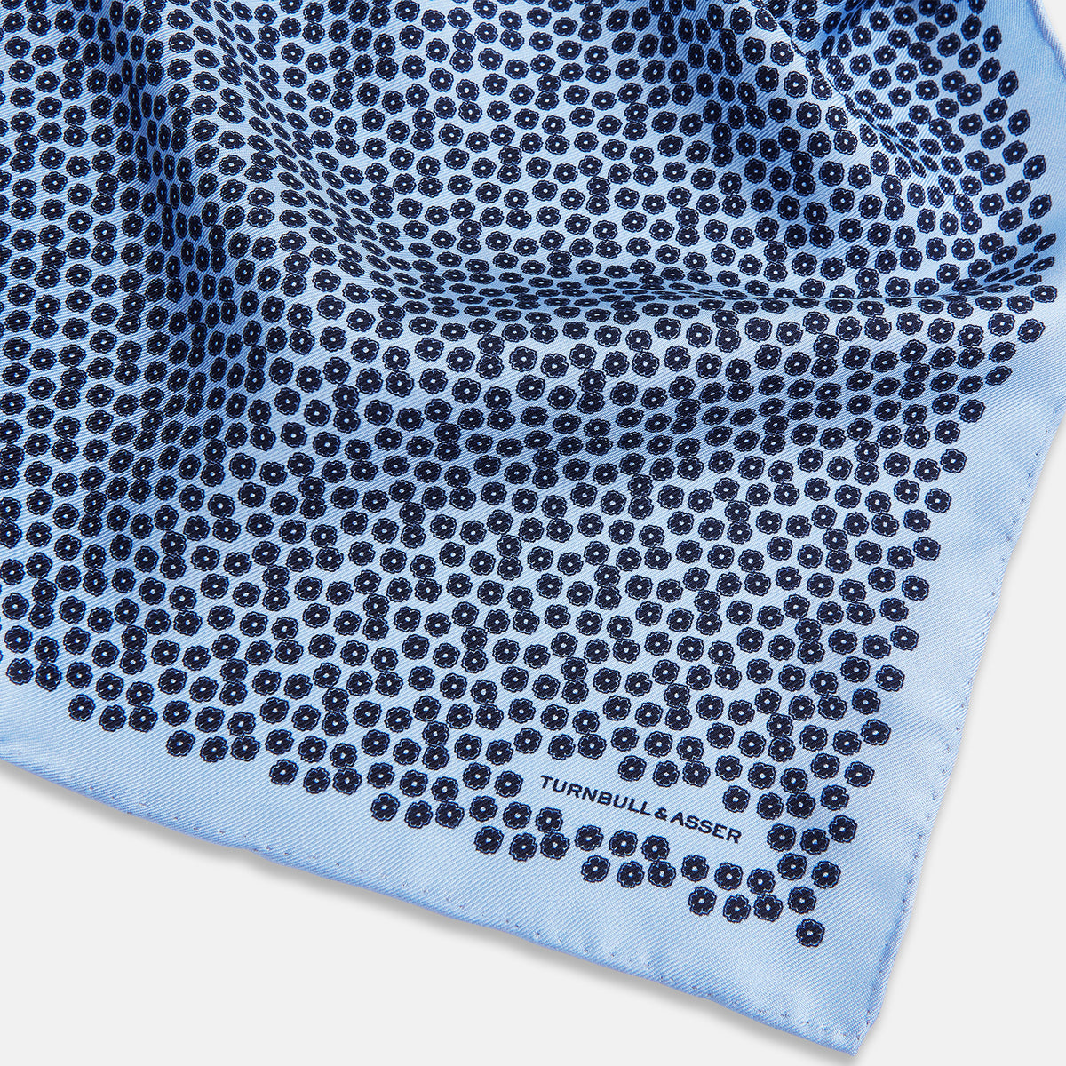 View 2 of product A light blue double-sided pocket square made of silk, featuring a dense pattern of small, dark blue floral motifs. One corner is folded over to reveal the Turnbull & Asser UK brand name printed in blue.