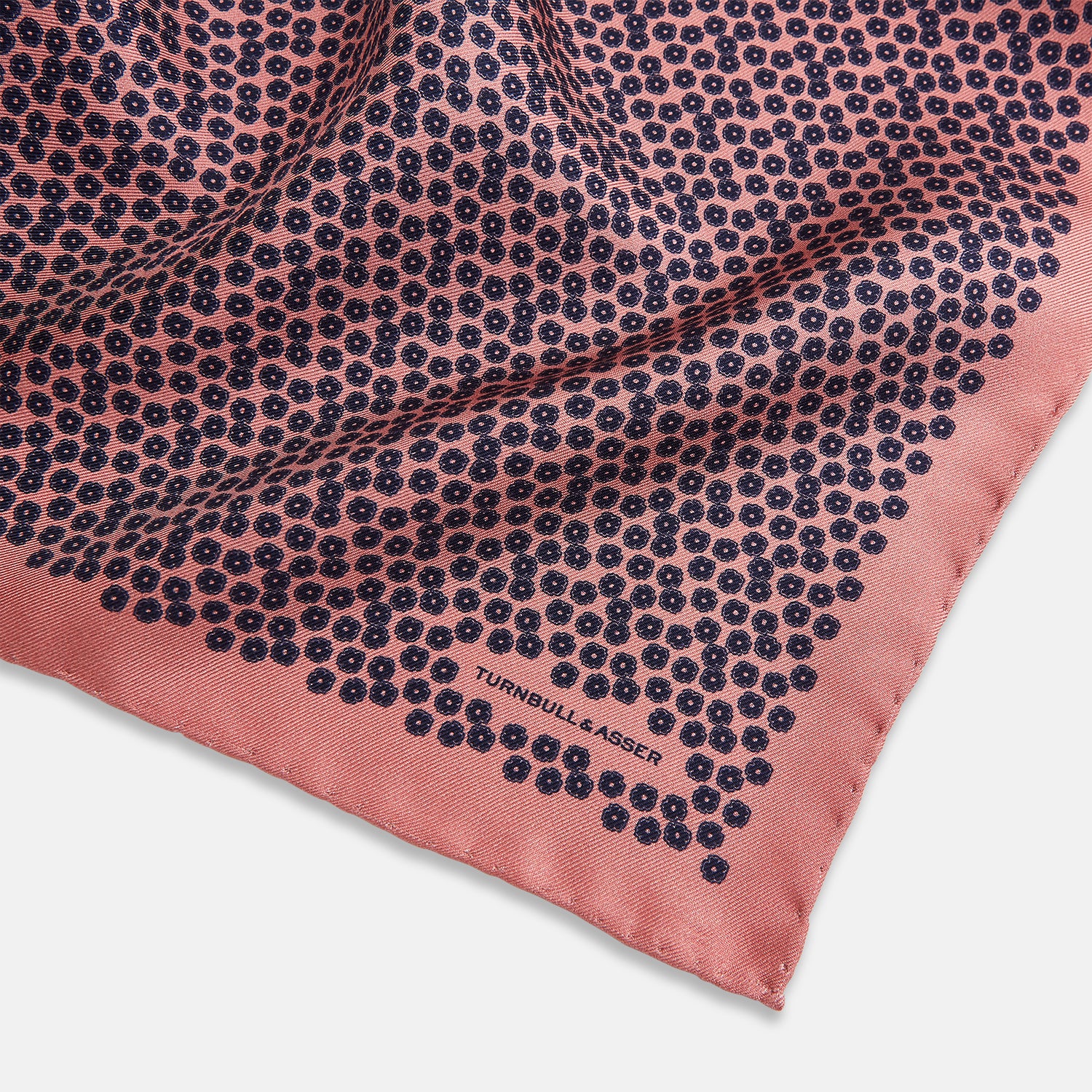 View 2 of product A folded Floral Pink Pocket Square in Silk by Turnbull & Asser UK, adorned with a pattern of small navy blue circles and the brand name printed in one corner, features a refined double-sided design for added versatility.