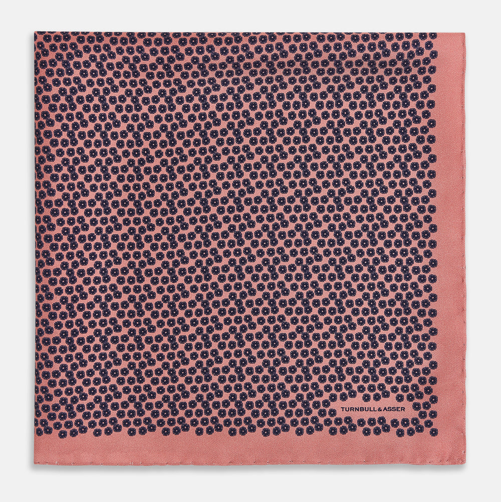 A folded Floral Pink Pocket Square in Silk by Turnbull & Asser UK, adorned with a pattern of small navy blue circles and the brand name printed in one corner, features a refined double-sided design for added versatility.