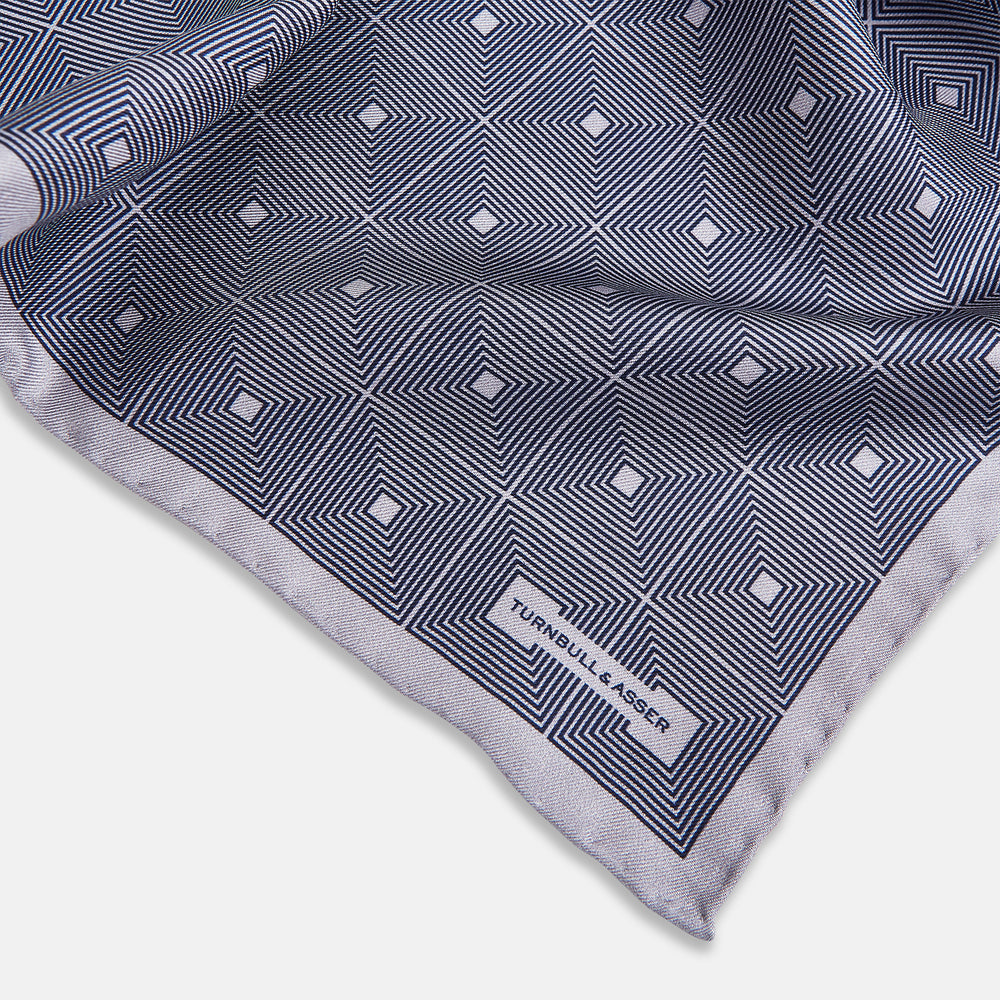 A folded, square Turnbull & Asser UK Geometric Grey Pocket Square in Silk featuring a geometric black and white concentric squares pattern accented with subtle navy lines, finished with a small Turnbull & Asser label in one corner.