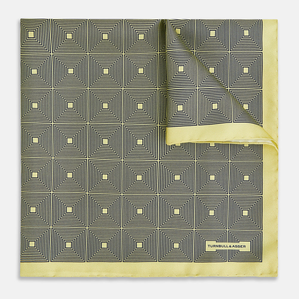 A checked yellow silk pocket square from Turnbull & Asser UK, featuring black and white concentric square designs with a yellow border. It is partially folded to highlight its double-sided pattern, and a small label reading TURNBULL & ASSER is visible in one corner.