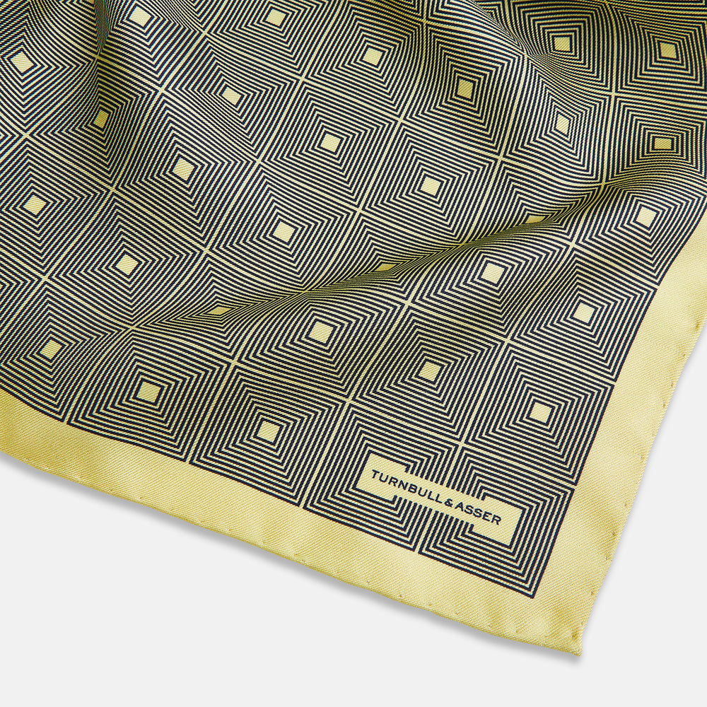 A checked yellow silk pocket square from Turnbull & Asser UK, featuring black and white concentric square designs with a yellow border. It is partially folded to highlight its double-sided pattern, and a small label reading TURNBULL & ASSER is visible in one corner.