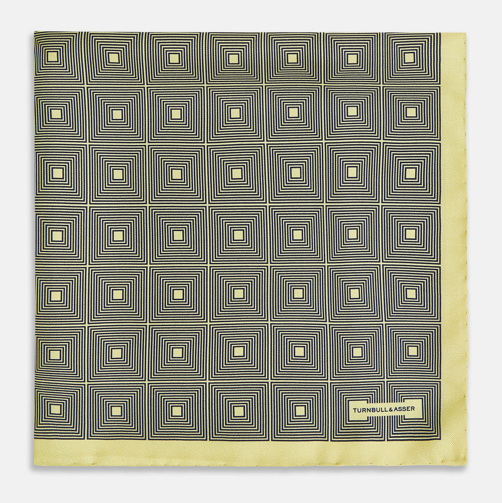 A checked yellow silk pocket square from Turnbull & Asser UK, featuring black and white concentric square designs with a yellow border. It is partially folded to highlight its double-sided pattern, and a small label reading TURNBULL & ASSER is visible in one corner.