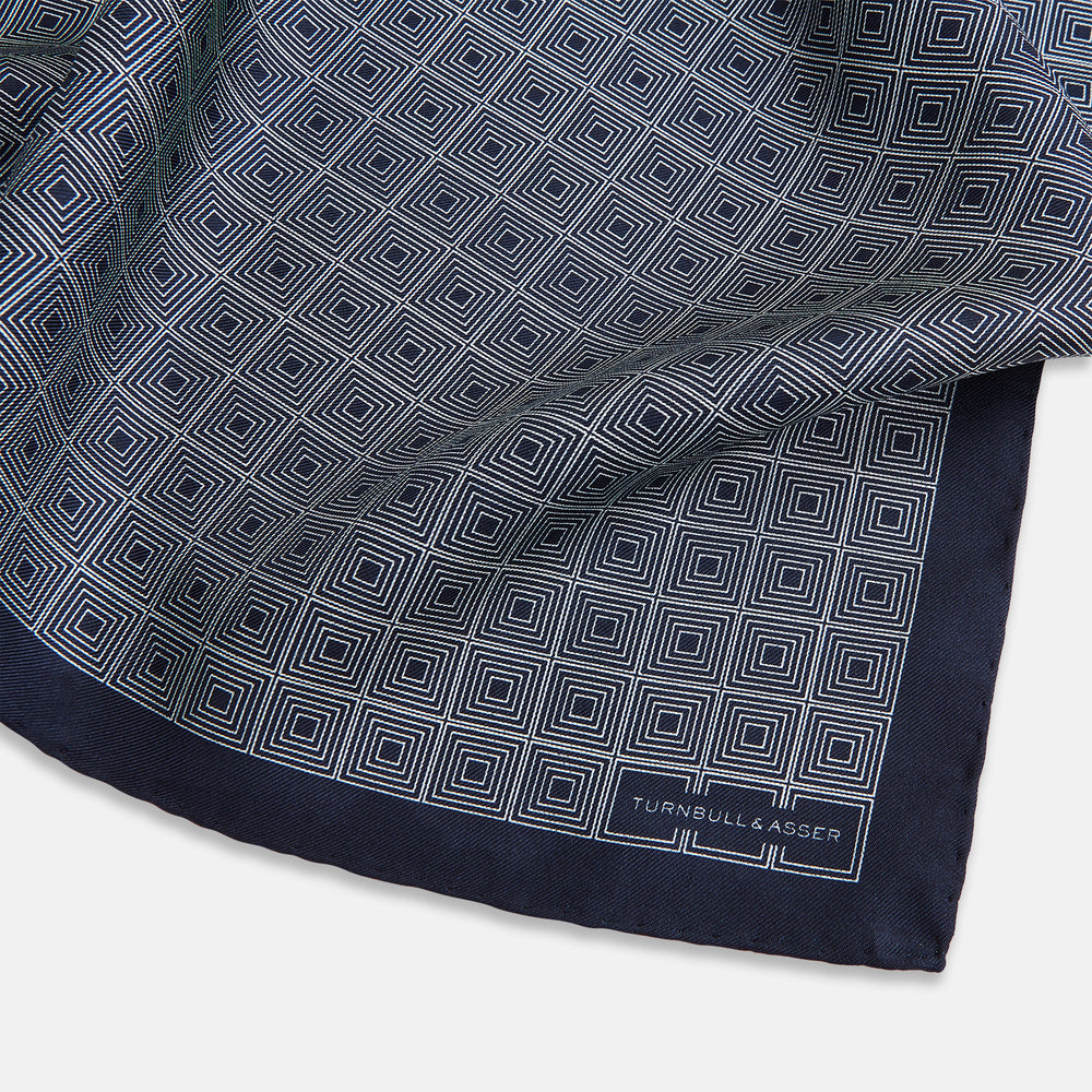 A Geometric Navy Pocket Square in Silk by Turnbull & Asser UK, showcasing a geometric pattern of small, overlapping concentric white squares and a stitched border. The pocket square is pictured with one corner folded over to display the Turnbull & Asser UK brand label.