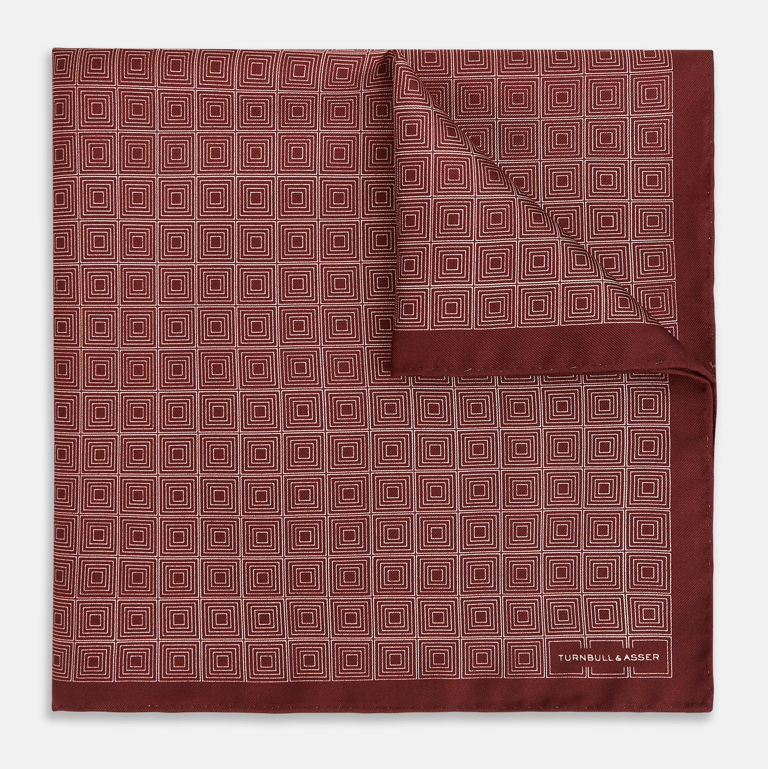 View 1 of product The Geometric Burgundy Pocket Square in Silk by Turnbull & Asser UK features a refined pattern of small, repeating white squares on a rich burgundy background—an ideal accessory for introducing sophistication to any outfit. One corner is artfully folded over to showcase the reverse side of this elegant geometric design, and the "Turnbull & Asser" label appears in the bottom right corner.
