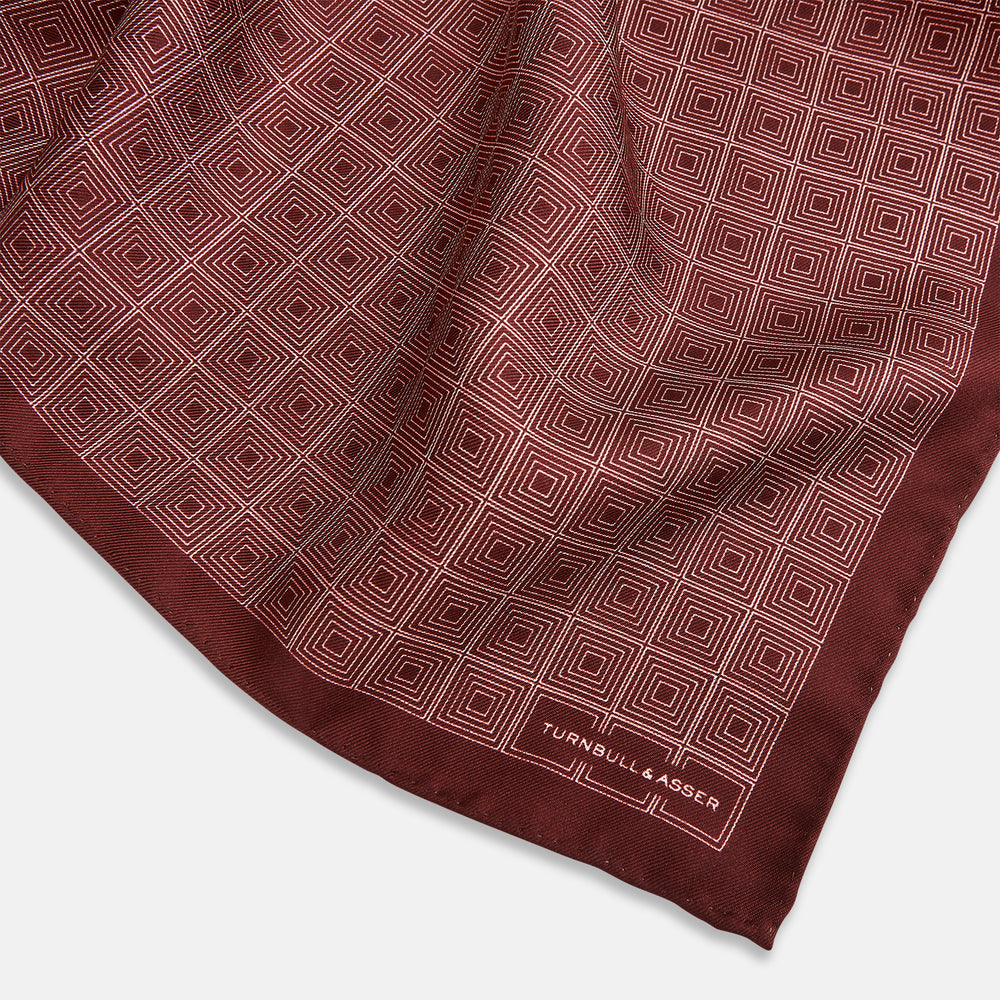 The Geometric Burgundy Pocket Square in Silk by Turnbull & Asser UK features a refined pattern of small, repeating white squares on a rich burgundy background—an ideal accessory for introducing sophistication to any outfit. One corner is artfully folded over to showcase the reverse side of this elegant geometric design, and the "Turnbull & Asser" label appears in the bottom right corner.