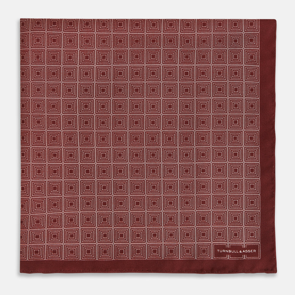 The Geometric Burgundy Pocket Square in Silk by Turnbull & Asser UK features a refined pattern of small, repeating white squares on a rich burgundy background—an ideal accessory for introducing sophistication to any outfit. One corner is artfully folded over to showcase the reverse side of this elegant geometric design, and the "Turnbull & Asser" label appears in the bottom right corner.