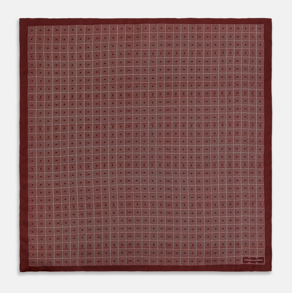 The Geometric Burgundy Pocket Square in Silk by Turnbull & Asser UK features a refined pattern of small, repeating white squares on a rich burgundy background—an ideal accessory for introducing sophistication to any outfit. One corner is artfully folded over to showcase the reverse side of this elegant geometric design, and the "Turnbull & Asser" label appears in the bottom right corner.