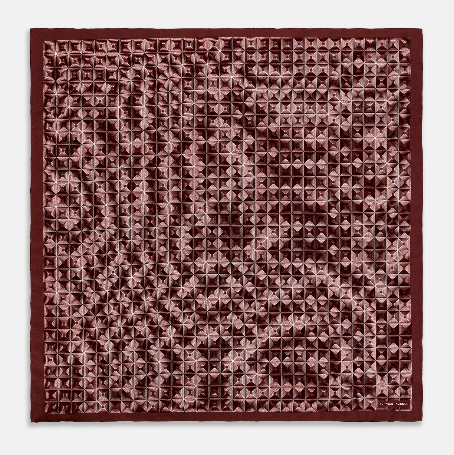 View 4 of product The Geometric Burgundy Pocket Square in Silk by Turnbull & Asser UK features a refined pattern of small, repeating white squares on a rich burgundy background—an ideal accessory for introducing sophistication to any outfit. One corner is artfully folded over to showcase the reverse side of this elegant geometric design, and the "Turnbull & Asser" label appears in the bottom right corner.