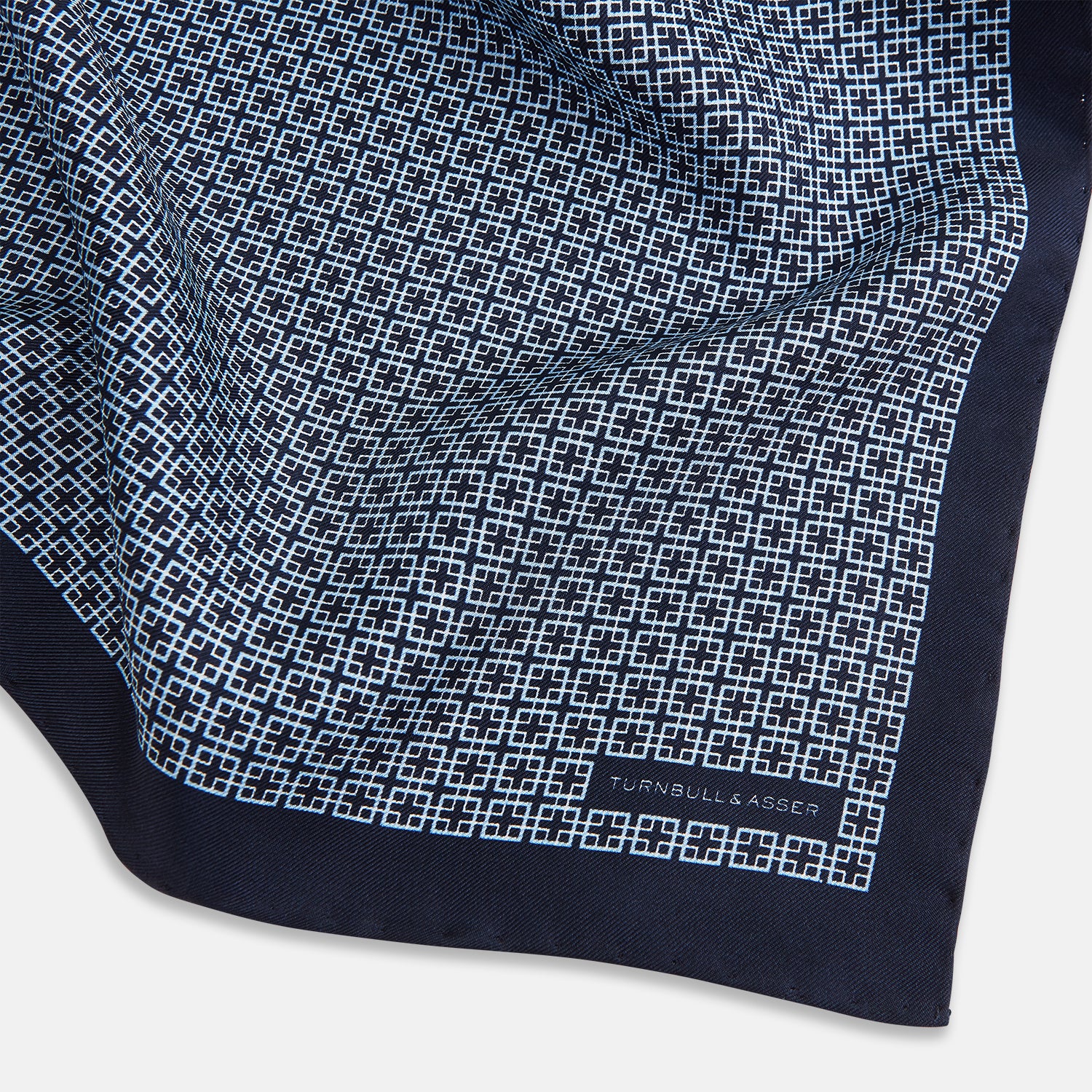 View 2 of product Introducing the Geometric Navy Pocket Square in Silk by Turnbull & Asser UK. This sophisticated accessory showcases a small, repeating geometric pattern in light blue and white on a rich navy background. The pocket square features a solid navy border, while the folded corner reveals the underside adorned with the Turnbull & Asser logo—an elegant finishing touch for any formal ensemble.