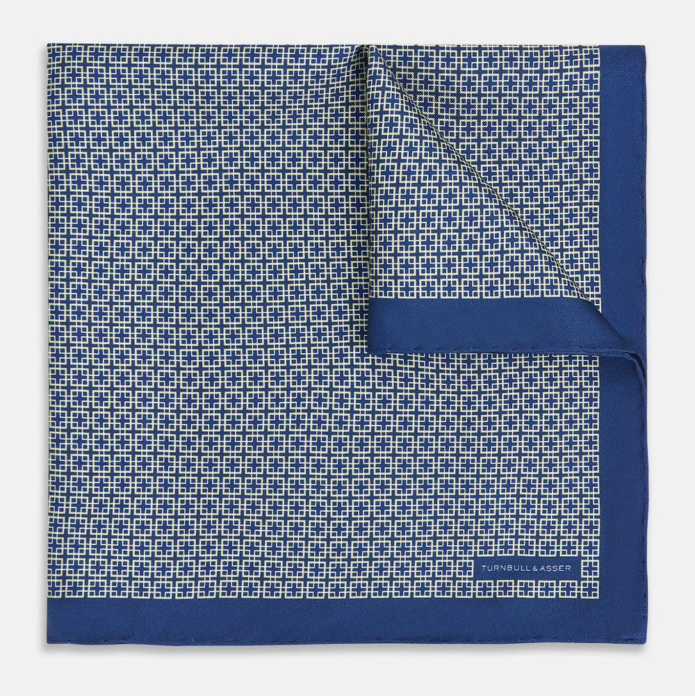 A blue pocket square from Turnbull & Asser UK, featuring a geometric white square pattern and a solid blue border. One corner is folded to highlight the striking design on both sides, with the brand name displayed on the bottom right. Product name: POCKET SQUARE SQUARE PATTERNED BANANA.