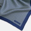 View 2 of product A blue pocket square from Turnbull & Asser UK, featuring a geometric white square pattern and a solid blue border. One corner is folded to highlight the striking design on both sides, with the brand name displayed on the bottom right. Product name: POCKET SQUARE SQUARE PATTERNED BANANA.