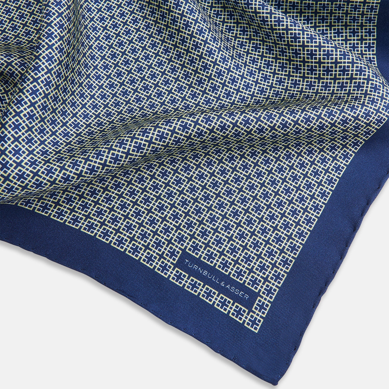 View 2 of product A blue pocket square from Turnbull & Asser UK, featuring a geometric white square pattern and a solid blue border. One corner is folded to highlight the striking design on both sides, with the brand name displayed on the bottom right. Product name: POCKET SQUARE SQUARE PATTERNED BANANA.