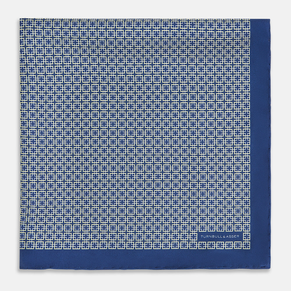 A blue pocket square from Turnbull & Asser UK, featuring a geometric white square pattern and a solid blue border. One corner is folded to highlight the striking design on both sides, with the brand name displayed on the bottom right. Product name: POCKET SQUARE SQUARE PATTERNED BANANA.