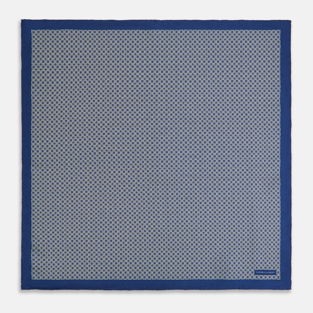 A blue pocket square from Turnbull & Asser UK, featuring a geometric white square pattern and a solid blue border. One corner is folded to highlight the striking design on both sides, with the brand name displayed on the bottom right. Product name: POCKET SQUARE SQUARE PATTERNED BANANA.