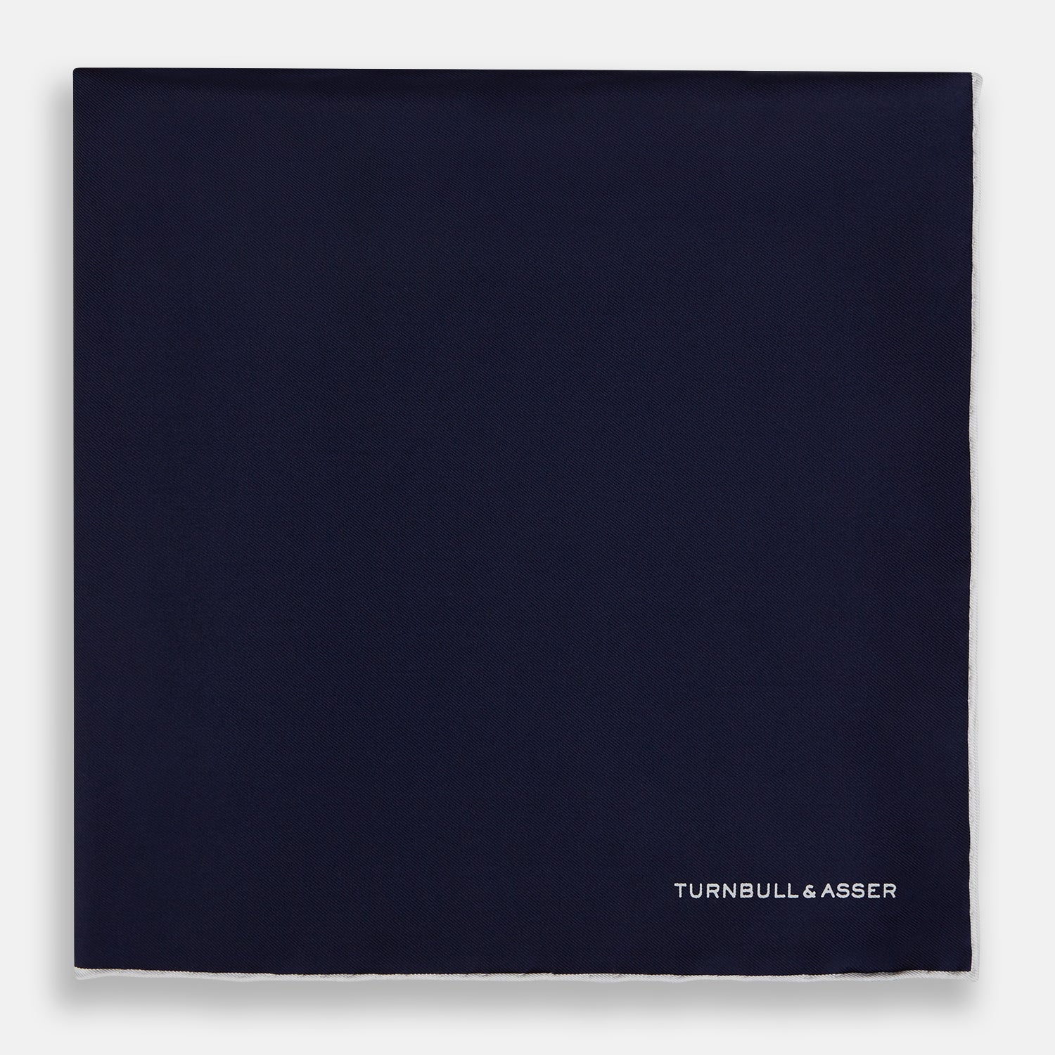 View 3 of product The Navy Piped Silk Pocket Square by Turnbull & Asser features white-stitched edges and subtle brand embroidery, ideal for enhancing formal looks. Shown neatly folded on a plain white background.