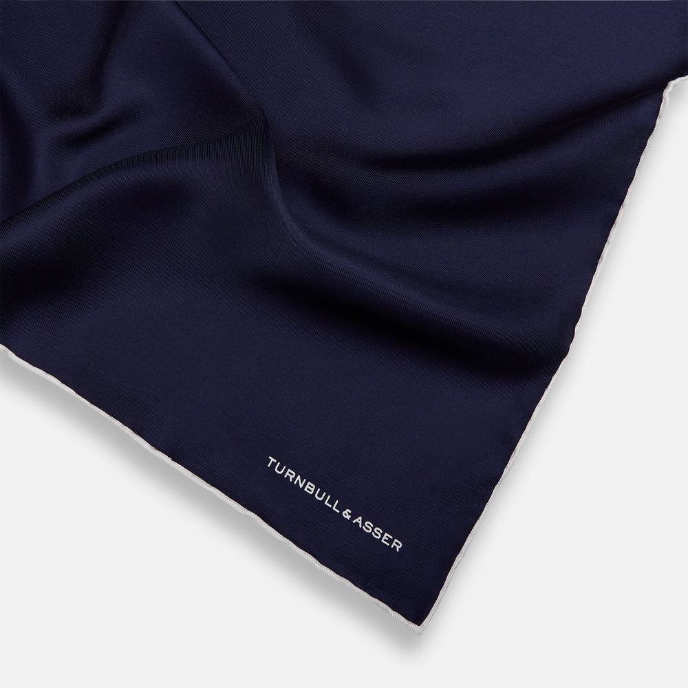 The Navy Piped Silk Pocket Square by Turnbull & Asser features white-stitched edges and subtle brand embroidery, ideal for enhancing formal looks. Shown neatly folded on a plain white background.