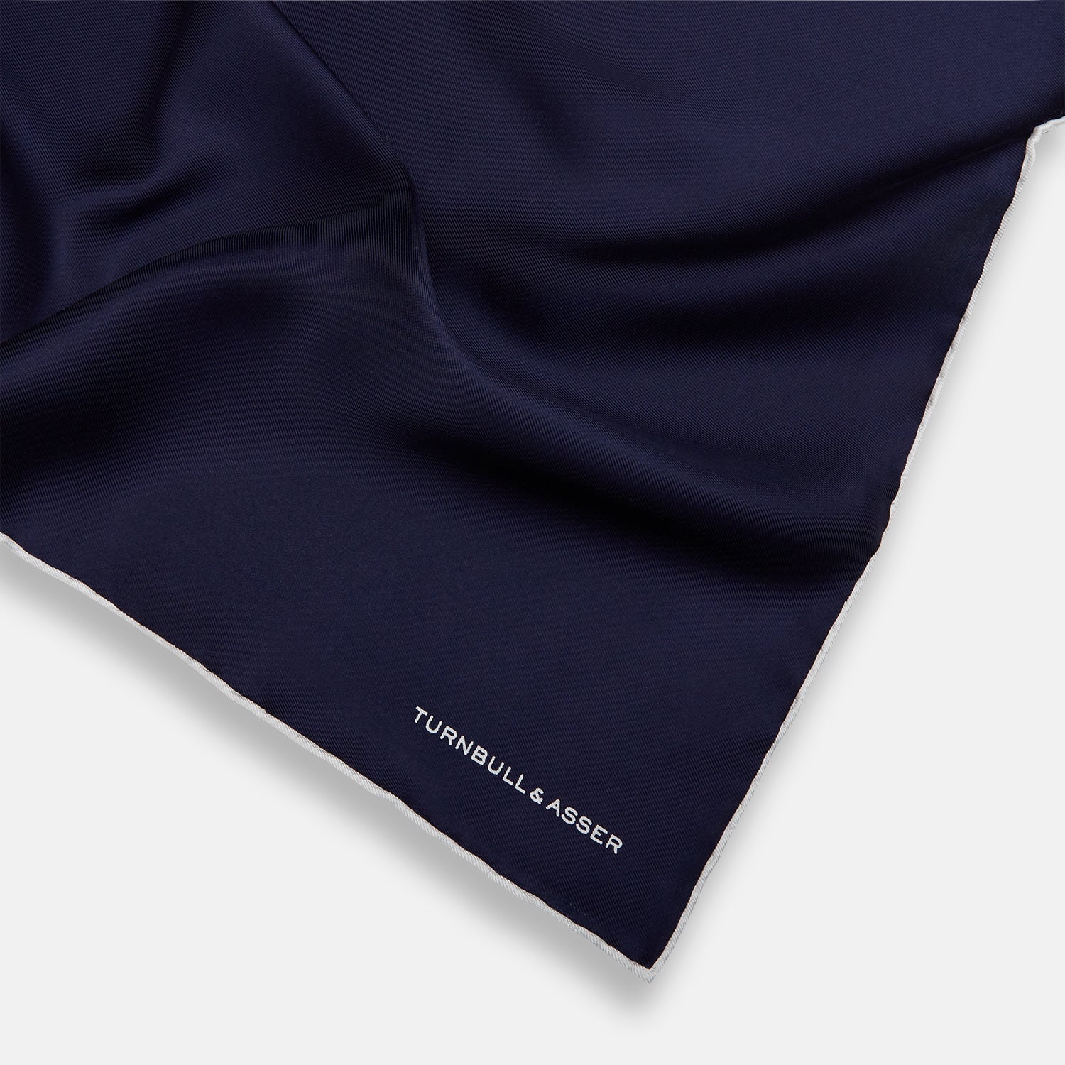 View 2 of product The Navy Piped Silk Pocket Square by Turnbull & Asser features white-stitched edges and subtle brand embroidery, ideal for enhancing formal looks. Shown neatly folded on a plain white background.
