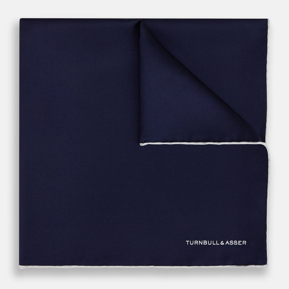 The Navy Piped Silk Pocket Square by Turnbull & Asser features white-stitched edges and subtle brand embroidery, ideal for enhancing formal looks. Shown neatly folded on a plain white background.