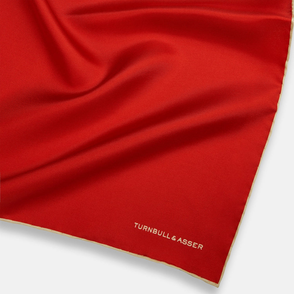 The Turnbull & Asser RED AND GOLD PIPED SILK POCKET SQUARE is a vibrant red silk accessory with gold edging and the brand’s logo in white on the bottom right—ideal for elevating formal looks.