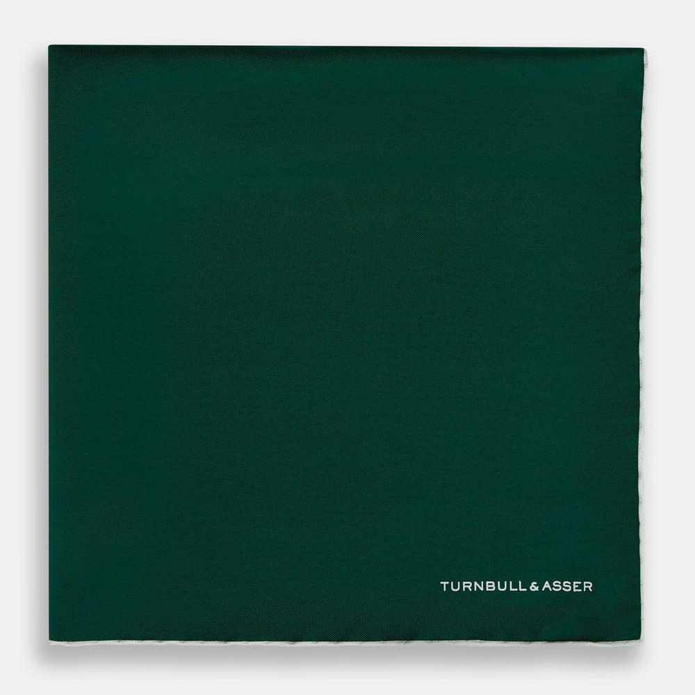 The FOREST GREEN AND ECRU PIPED SILK POCKET SQUARE by Turnbull & Asser features forest green silk with ecru piping, neatly folded to display both sides, and the brand name printed in white in the lower right corner—ideal for formal attire.