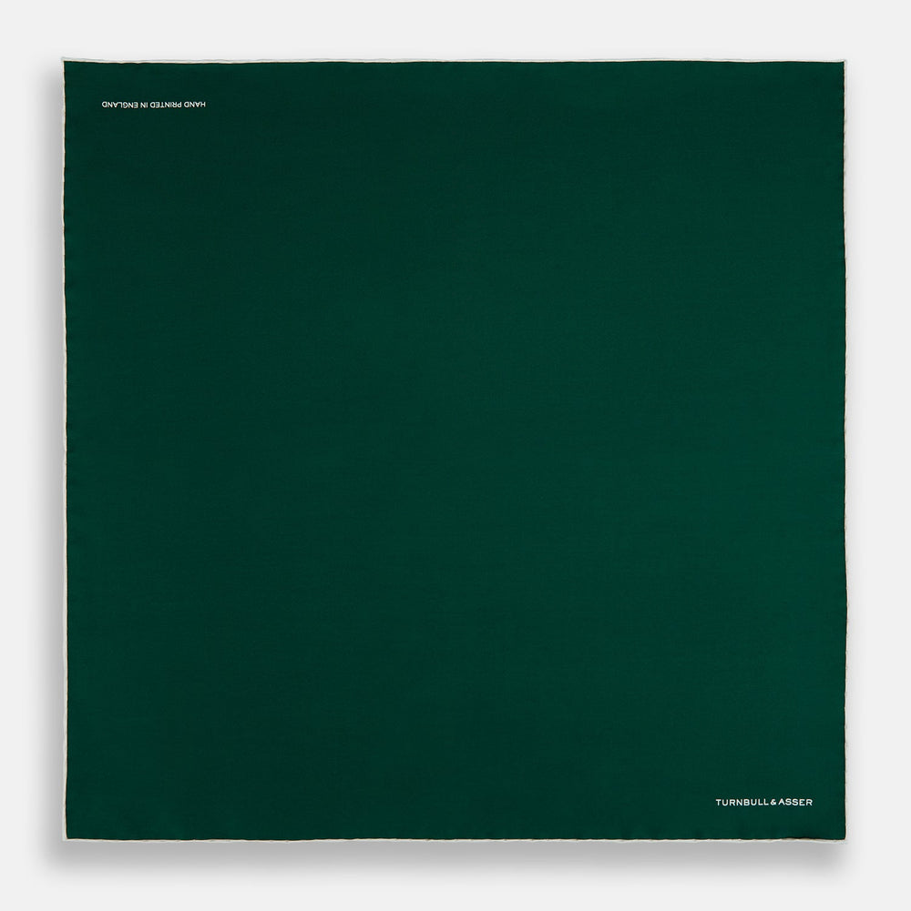 The FOREST GREEN AND ECRU PIPED SILK POCKET SQUARE by Turnbull & Asser features forest green silk with ecru piping, neatly folded to display both sides, and the brand name printed in white in the lower right corner—ideal for formal attire.