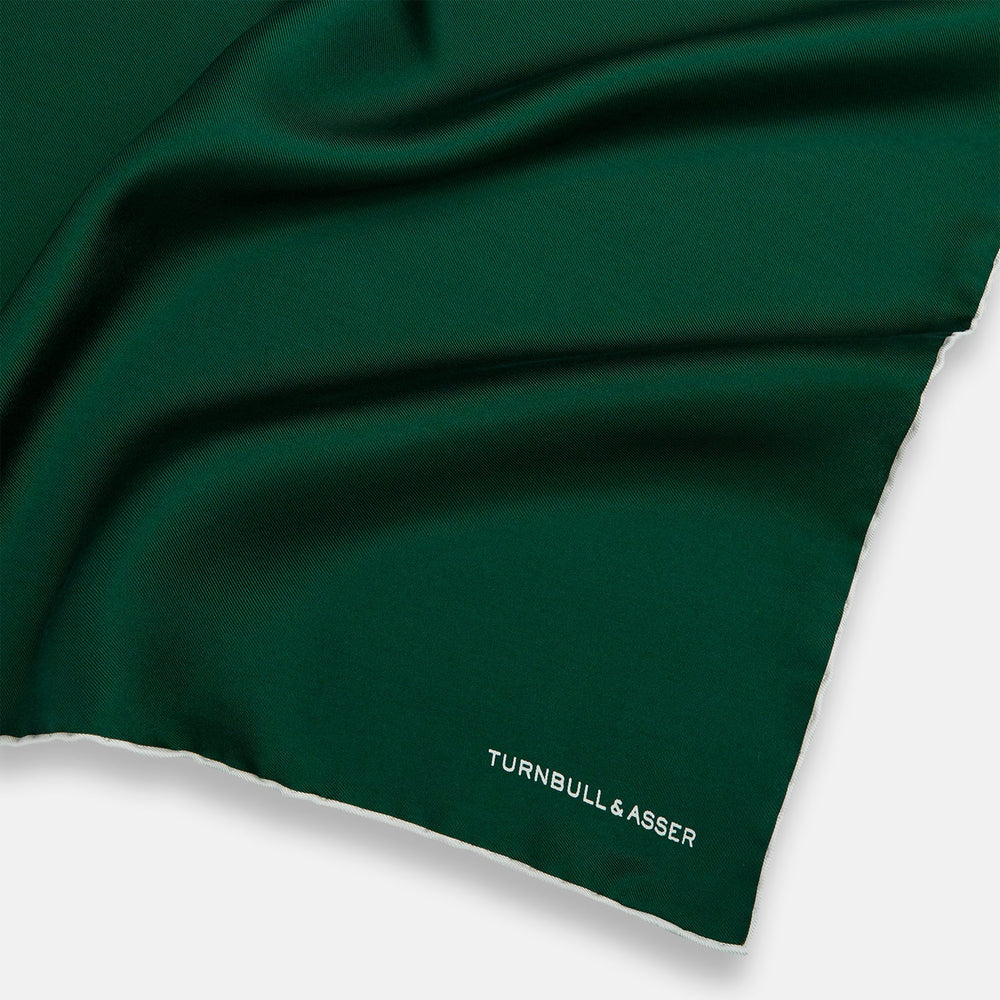 The FOREST GREEN AND ECRU PIPED SILK POCKET SQUARE by Turnbull & Asser features forest green silk with ecru piping, neatly folded to display both sides, and the brand name printed in white in the lower right corner—ideal for formal attire.