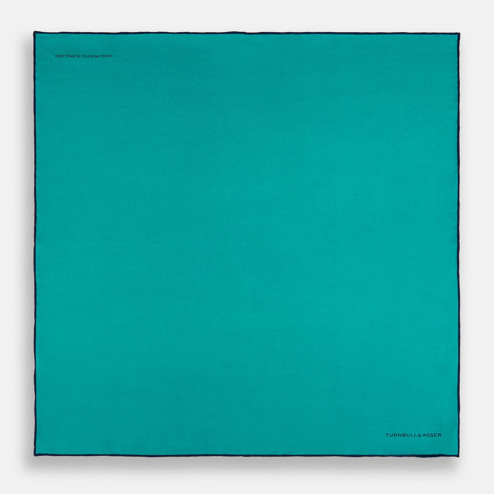 The Turquoise Piped Silk Pocket Square by Turnbull & Asser features dark blue edging and the brand’s logo, offering a sophisticated accent to formal outfits.