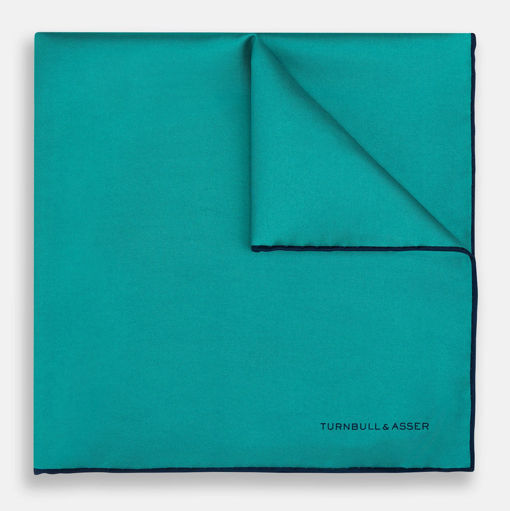 The Turquoise Piped Silk Pocket Square by Turnbull & Asser features dark blue edging and the brand’s logo, offering a sophisticated accent to formal outfits.