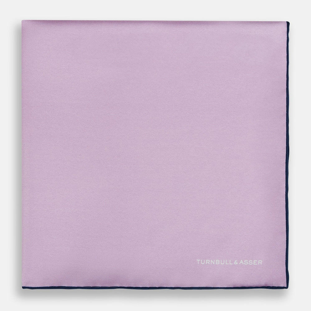 The Turnbull & Asser Lilac Piped Silk Pocket Square features dark edging and a white brand logo in one corner, making it a stylish choice for formal attire. Displayed against a white background.