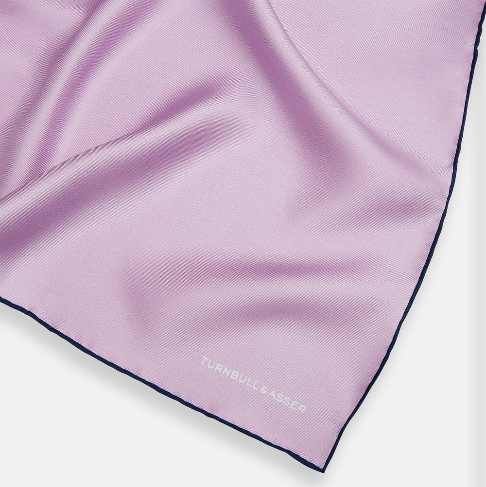 The Turnbull & Asser Lilac Piped Silk Pocket Square features dark edging and a white brand logo in one corner, making it a stylish choice for formal attire. Displayed against a white background.