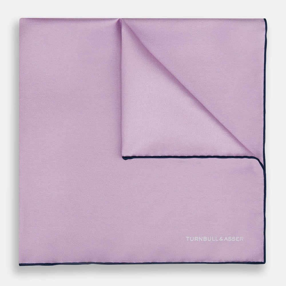 The Turnbull & Asser Lilac Piped Silk Pocket Square features dark edging and a white brand logo in one corner, making it a stylish choice for formal attire. Displayed against a white background.