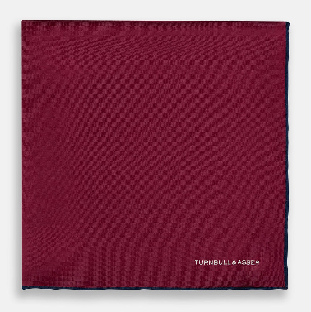 The Mauve Piped Silk Pocket Square by Turnbull & Asser features a dark blue edge and white embroidered logo in the lower right corner, making it ideal for formal occasions. Displayed on a plain white background.
