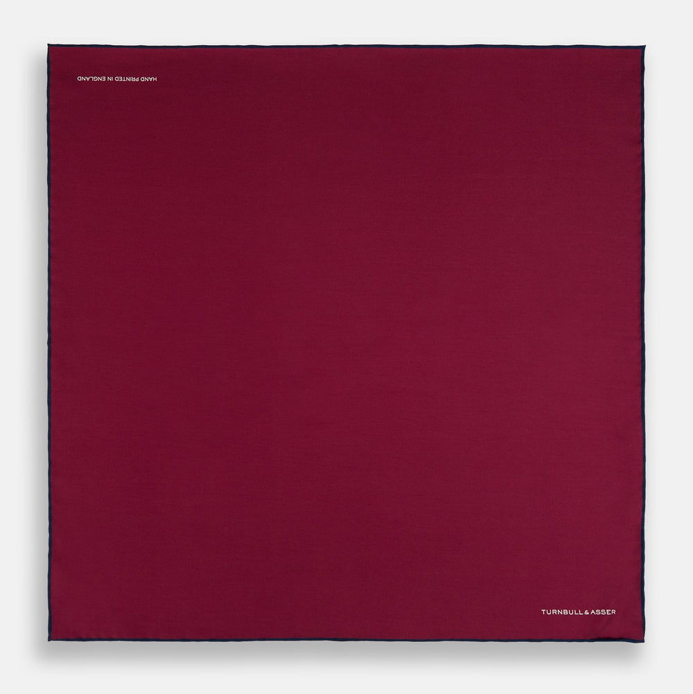 The Mauve Piped Silk Pocket Square by Turnbull & Asser features a dark blue edge and white embroidered logo in the lower right corner, making it ideal for formal occasions. Displayed on a plain white background.