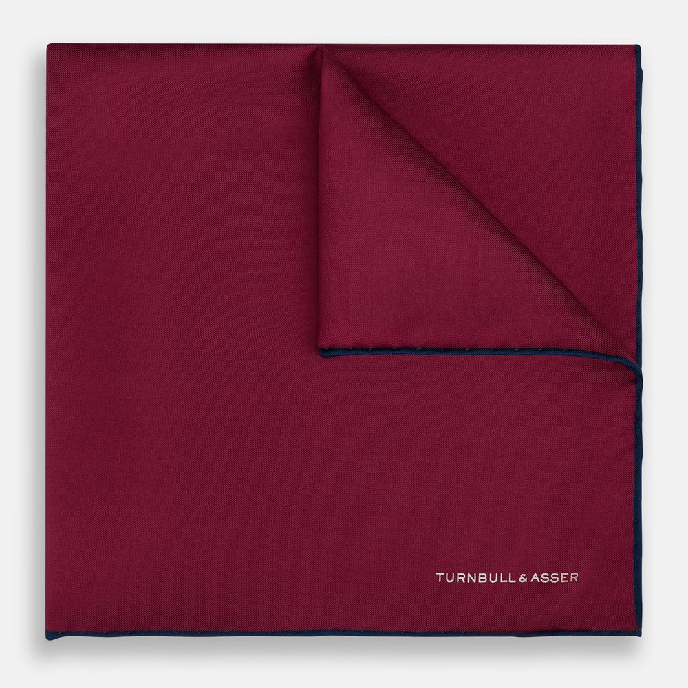 The Mauve Piped Silk Pocket Square by Turnbull & Asser features a dark blue edge and white embroidered logo in the lower right corner, making it ideal for formal occasions. Displayed on a plain white background.