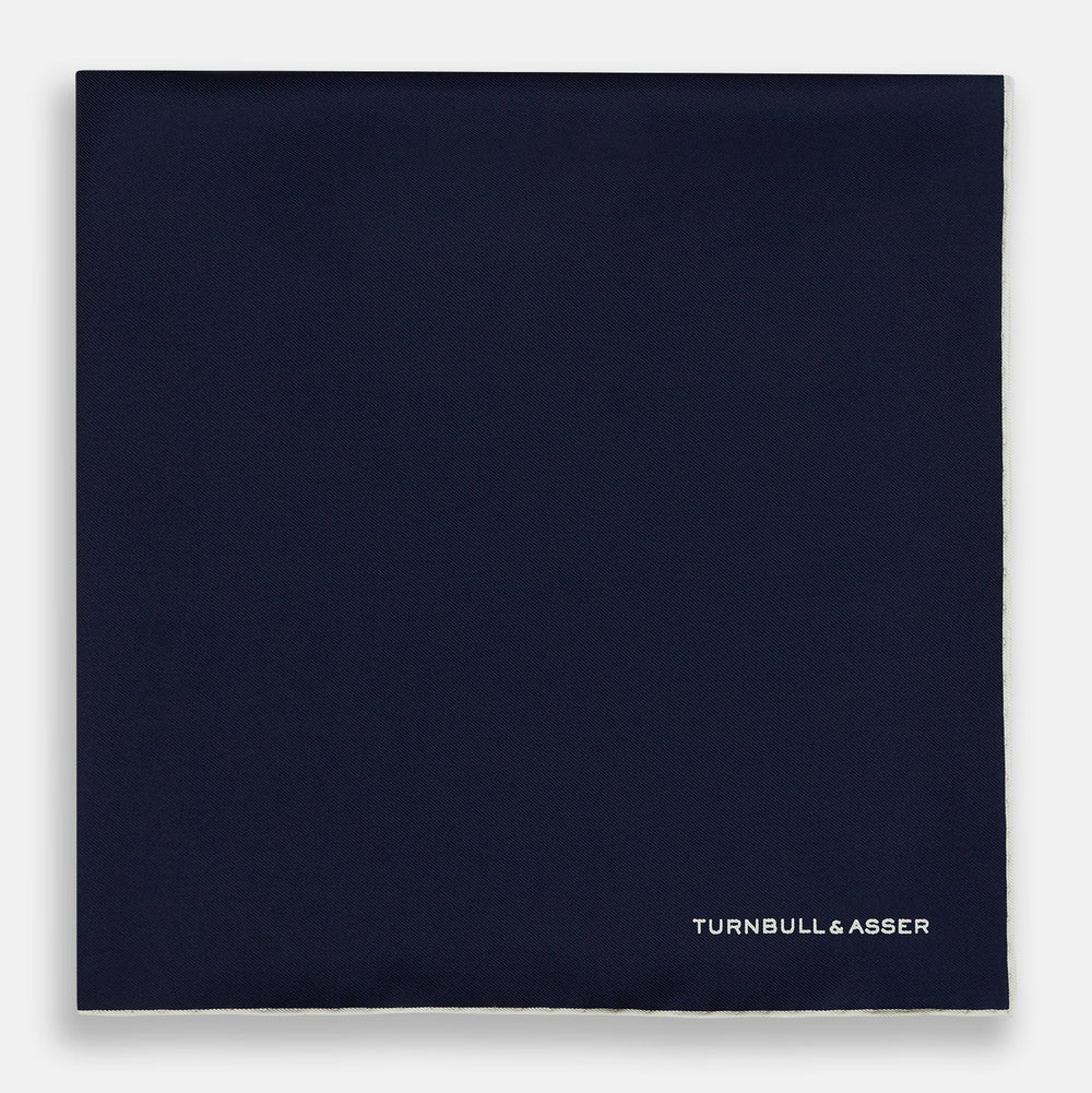 The Dark Blue Piped Silk Pocket Square by Turnbull & Asser features white piping, a one-corner fold revealing the underside, and the brand's name embroidered in white—perfect for formal occasions.