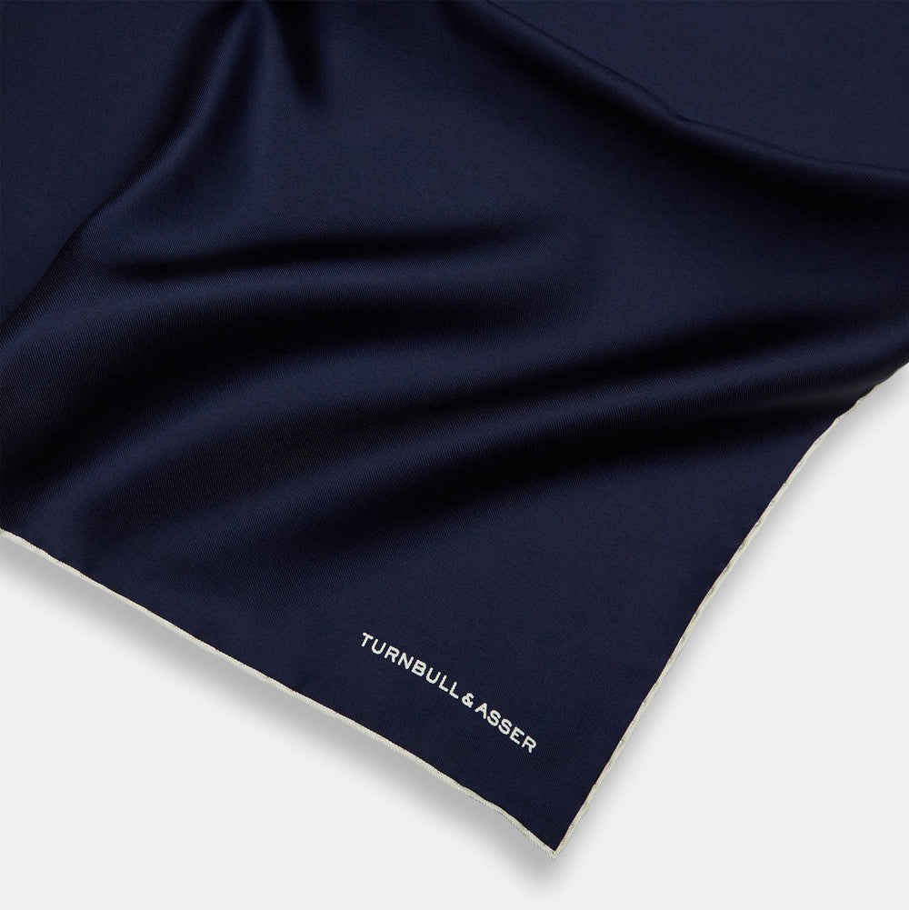 The Dark Blue Piped Silk Pocket Square by Turnbull & Asser features white piping, a one-corner fold revealing the underside, and the brand's name embroidered in white—perfect for formal occasions.