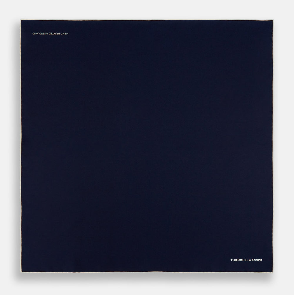 The Dark Blue Piped Silk Pocket Square by Turnbull & Asser features white piping, a one-corner fold revealing the underside, and the brand's name embroidered in white—perfect for formal occasions.