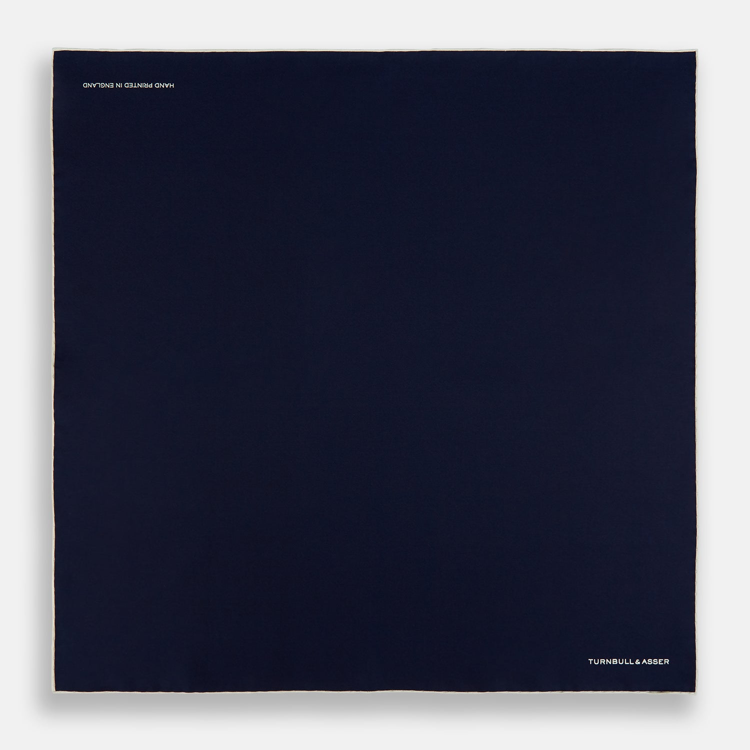View 4 of product The Dark Blue Piped Silk Pocket Square by Turnbull & Asser features white piping, a one-corner fold revealing the underside, and the brand's name embroidered in white—perfect for formal occasions.