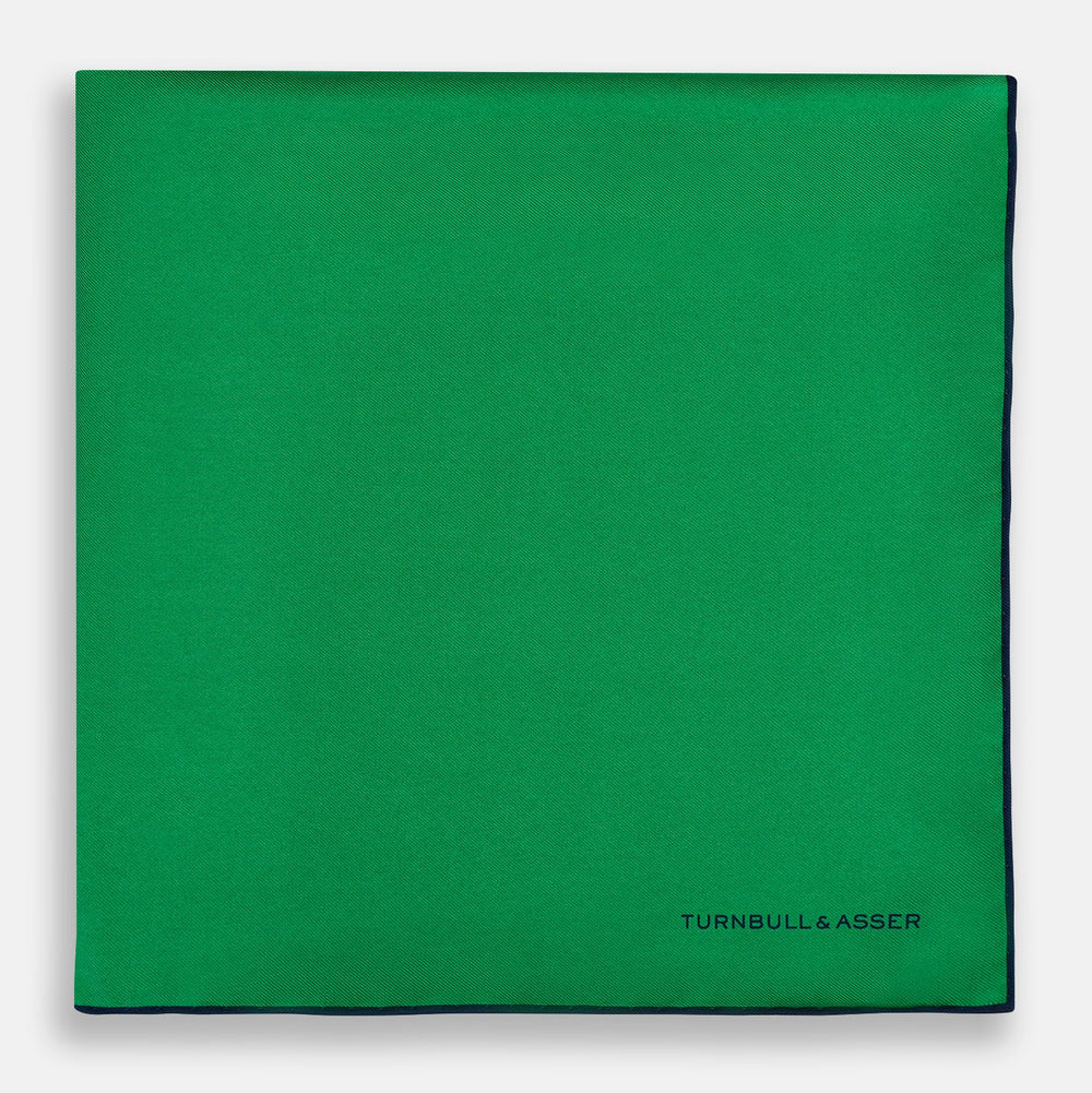 The Green Piped Silk Pocket Square by Turnbull & Asser features dark blue stitched edges and a neat fold—ideal for formal occasions. The Turnbull & Asser name is printed in black on the lower right corner.