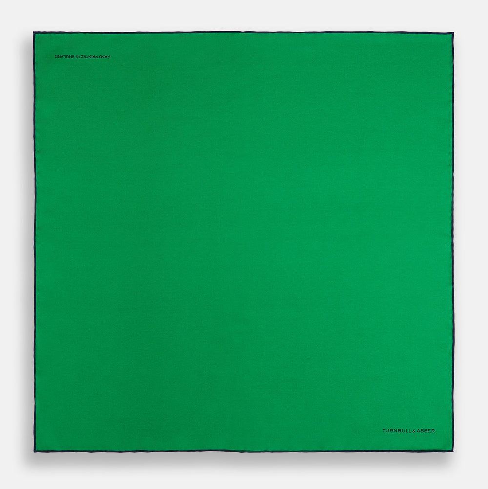 The Green Piped Silk Pocket Square by Turnbull & Asser features dark blue stitched edges and a neat fold—ideal for formal occasions. The Turnbull & Asser name is printed in black on the lower right corner.