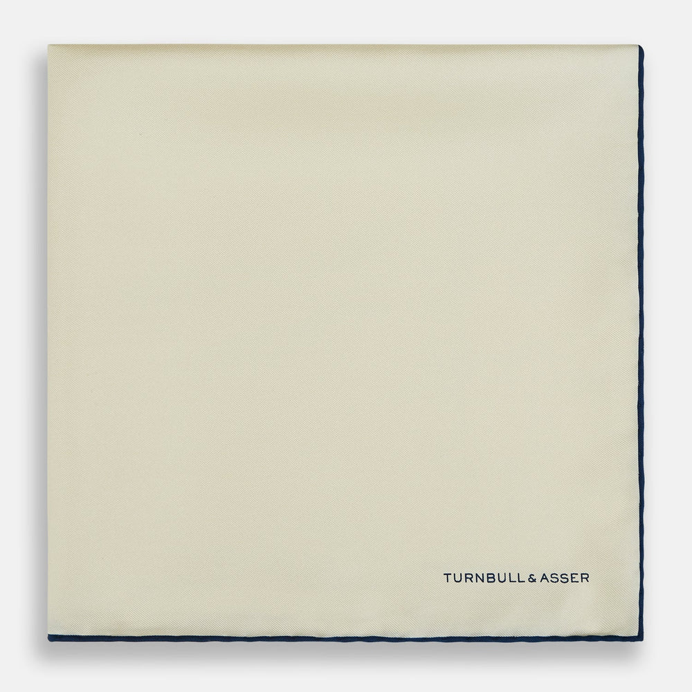 The Ecru and Navy Blue Piped Silk Pocket Square by Turnbull & Asser features contrasting navy piping and finely embroidered branding—an elegant finishing touch to any formal look.