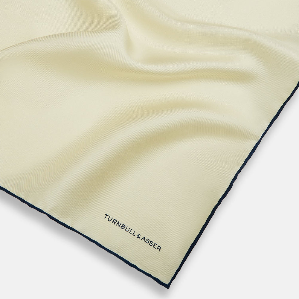 The Ecru and Navy Blue Piped Silk Pocket Square by Turnbull & Asser features contrasting navy piping and finely embroidered branding—an elegant finishing touch to any formal look.