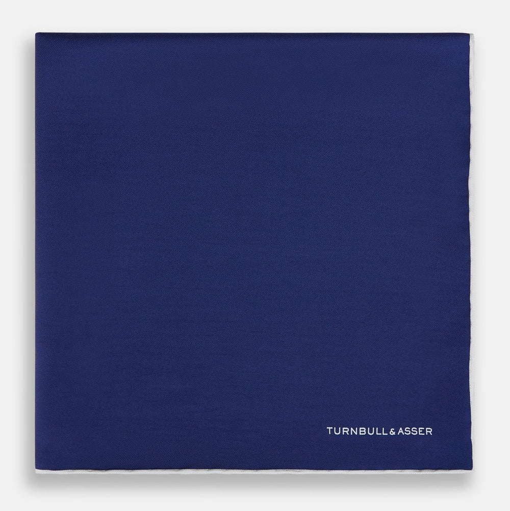 The Turnbull & Asser Mid Blue Piped Silk Pocket Square features a mid-blue hue with white edging, ideal for formal wear, and is finished with the brand’s name printed in white on the bottom right corner.