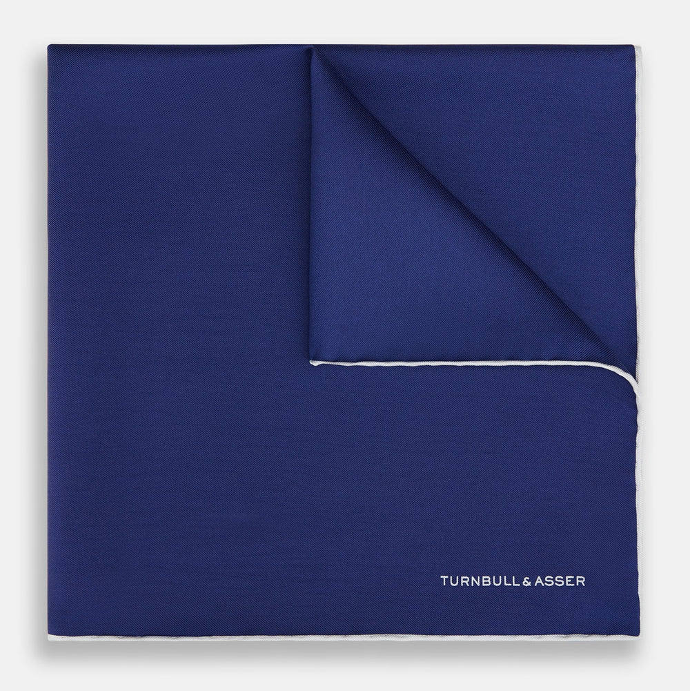 The Turnbull & Asser Mid Blue Piped Silk Pocket Square features a mid-blue hue with white edging, ideal for formal wear, and is finished with the brand’s name printed in white on the bottom right corner.