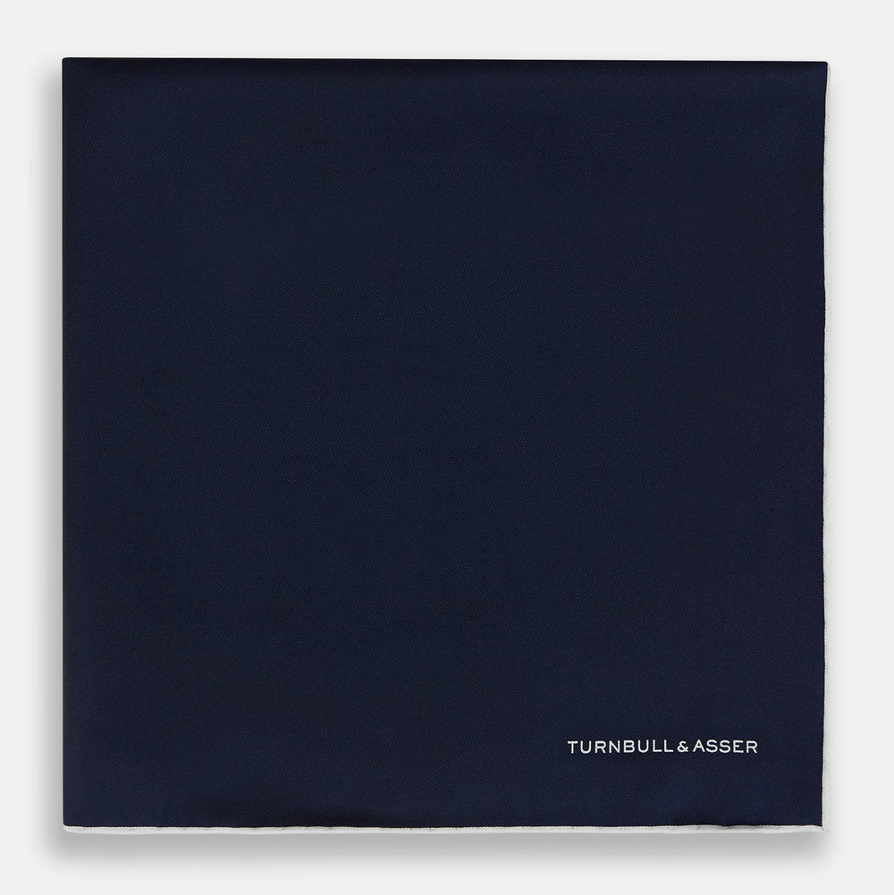The Midnight Blue Piped Silk Pocket Square from Turnbull & Asser features crisp white trim and subtle branding, making it the ideal accessory to elevate your formal look.