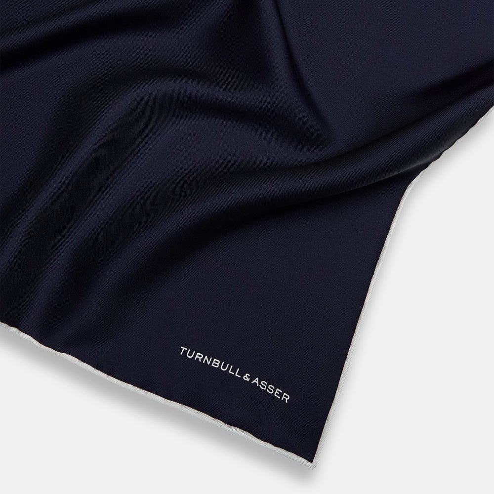 The Midnight Blue Piped Silk Pocket Square from Turnbull & Asser features crisp white trim and subtle branding, making it the ideal accessory to elevate your formal look.