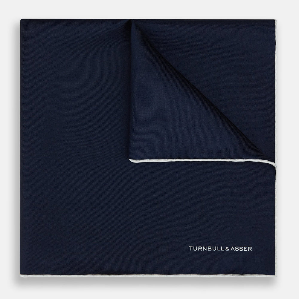The Midnight Blue Piped Silk Pocket Square from Turnbull & Asser features crisp white trim and subtle branding, making it the ideal accessory to elevate your formal look.